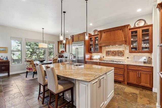 kitchen with appliances with stainless steel finishes, a sink, premium range hood, stone tile floors, and tasteful backsplash