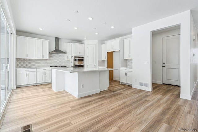 kitchen featuring tasteful backsplash, white cabinets, a kitchen island with sink, wall chimney range hood, and light wood-type flooring