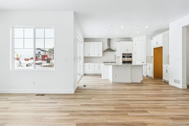 kitchen featuring double oven, an island with sink, white cabinets, backsplash, and wall chimney range hood