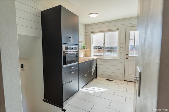 kitchen with wooden walls, light tile patterned floors, visible vents, stainless steel oven, and dark cabinets