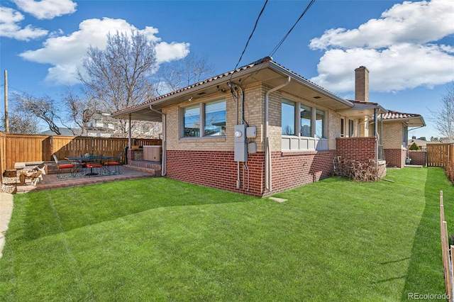 view of side of home with a patio, a fenced backyard, brick siding, a yard, and a chimney