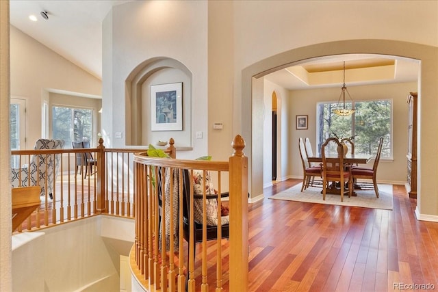 hall with baseboards, arched walkways, hardwood / wood-style floors, a tray ceiling, and an upstairs landing