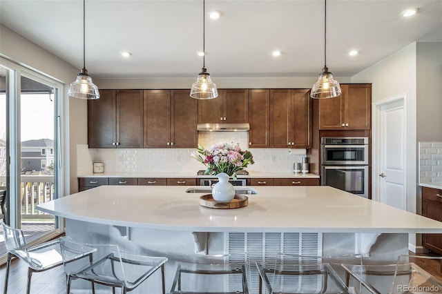 kitchen with decorative backsplash, stainless steel double oven, light countertops, under cabinet range hood, and an island with sink