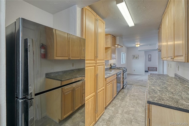 kitchen with light brown cabinetry, dark countertops, appliances with stainless steel finishes, wall chimney exhaust hood, and baseboards