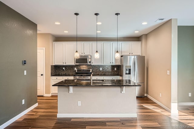 kitchen with stainless steel appliances, dark stone countertops, an island with sink, pendant lighting, and white cabinets