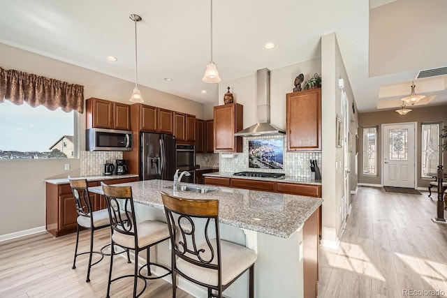 kitchen featuring appliances with stainless steel finishes, brown cabinetry, a sink, an island with sink, and wall chimney exhaust hood