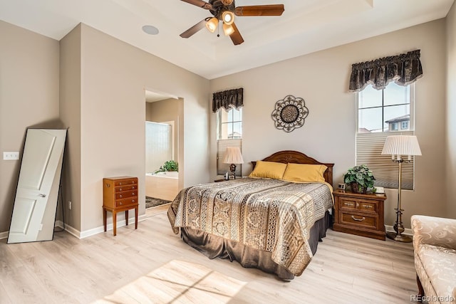 bedroom featuring a tray ceiling, light wood-style floors, ceiling fan, connected bathroom, and baseboards