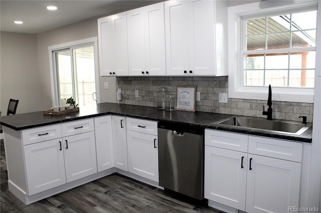 kitchen featuring dark countertops, stainless steel dishwasher, white cabinets, a sink, and a peninsula