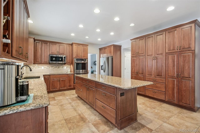 kitchen featuring stainless steel appliances, brown cabinetry, a kitchen island, a sink, and light stone countertops