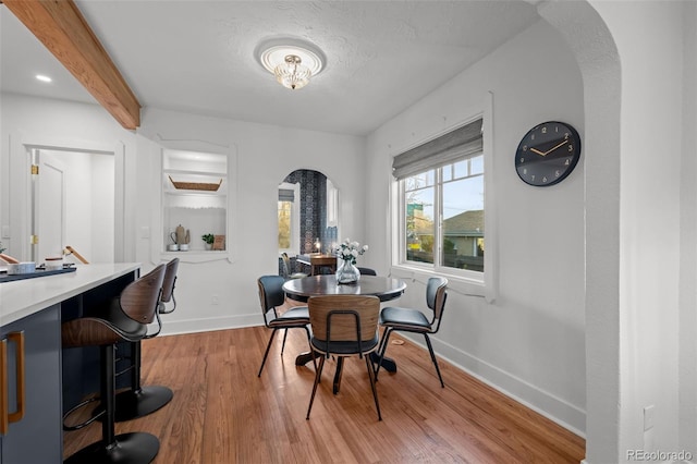dining space featuring arched walkways, baseboards, light wood-type flooring, and beam ceiling