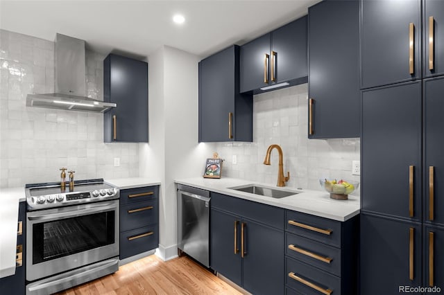 kitchen with wall chimney range hood, appliances with stainless steel finishes, a sink, light countertops, and blue cabinetry