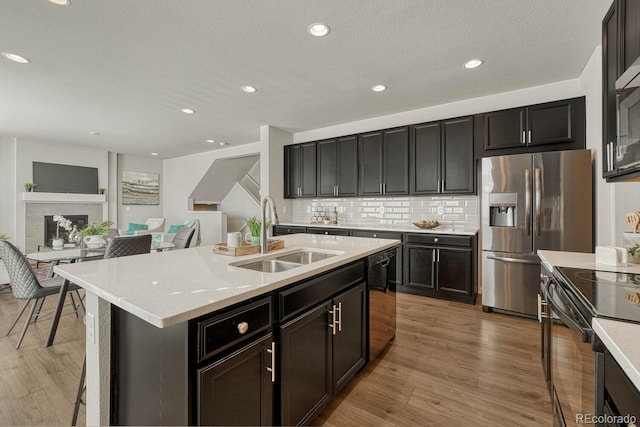 kitchen featuring sink, a tile fireplace, tasteful backsplash, a center island with sink, and appliances with stainless steel finishes