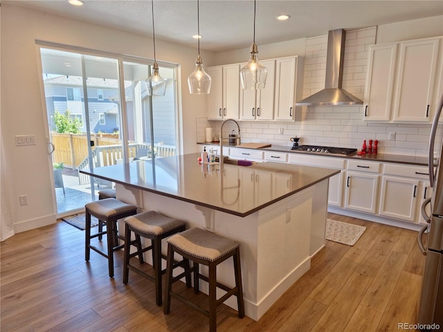 kitchen with a breakfast bar, white cabinets, wall chimney range hood, an island with sink, and light hardwood / wood-style floors