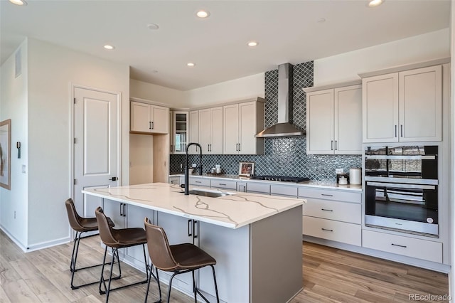 kitchen with sink, double wall oven, light stone countertops, a center island with sink, and wall chimney exhaust hood