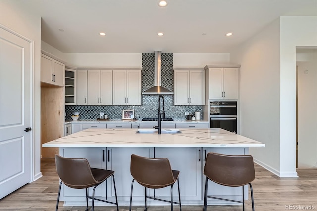 kitchen with double oven, decorative backsplash, a kitchen island with sink, light stone countertops, and wall chimney range hood