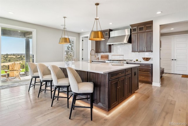 kitchen featuring wall chimney exhaust hood, sink, a center island with sink, pendant lighting, and light hardwood / wood-style floors