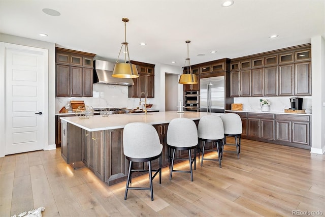 kitchen featuring appliances with stainless steel finishes, hanging light fixtures, a large island with sink, dark brown cabinetry, and wall chimney exhaust hood