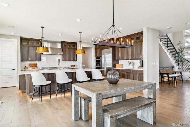 kitchen with decorative light fixtures, light hardwood / wood-style flooring, an island with sink, stainless steel appliances, and wall chimney range hood