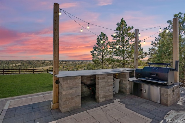 patio terrace at dusk with a yard, grilling area, and an outdoor kitchen