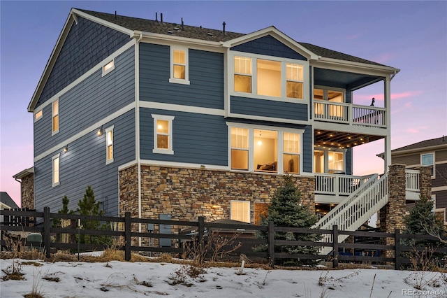 snow covered rear of property with a balcony, stone siding, and a fenced front yard