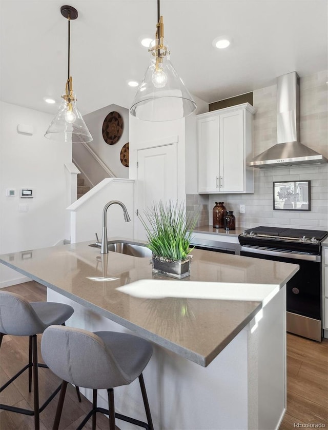 kitchen featuring stainless steel stove, an island with sink, white cabinets, decorative backsplash, and wall chimney range hood