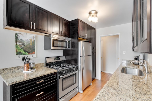 kitchen with dark brown cabinets, light wood-type flooring, light stone counters, a sink, and appliances with stainless steel finishes