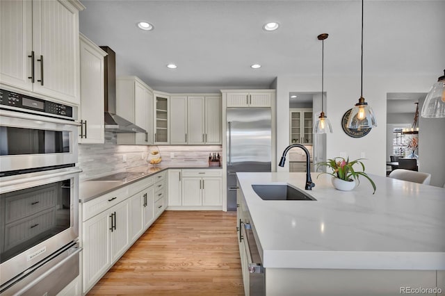 kitchen featuring decorative backsplash, a center island with sink, wall chimney exhaust hood, a sink, and appliances with stainless steel finishes