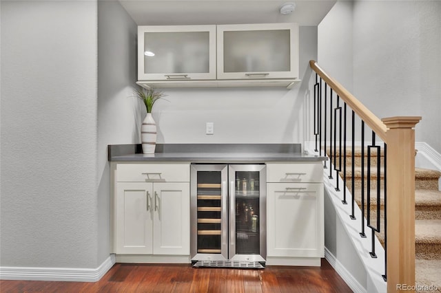 bar featuring dark wood-style flooring, beverage cooler, baseboards, a bar, and stairway
