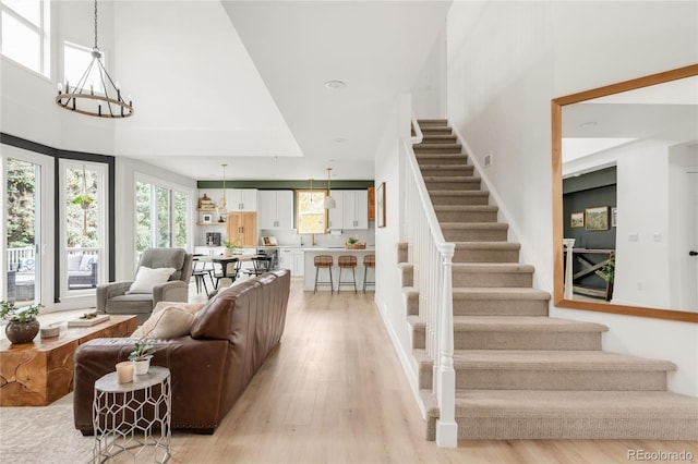 living area with stairs, an inviting chandelier, light wood-style flooring, and a towering ceiling