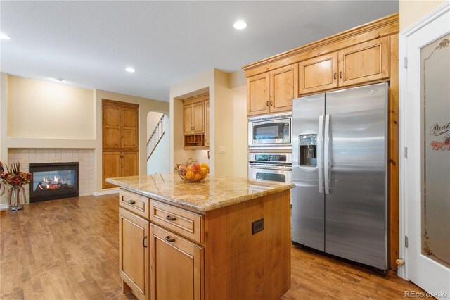 kitchen with a kitchen island, wood finished floors, appliances with stainless steel finishes, light stone countertops, and a tile fireplace