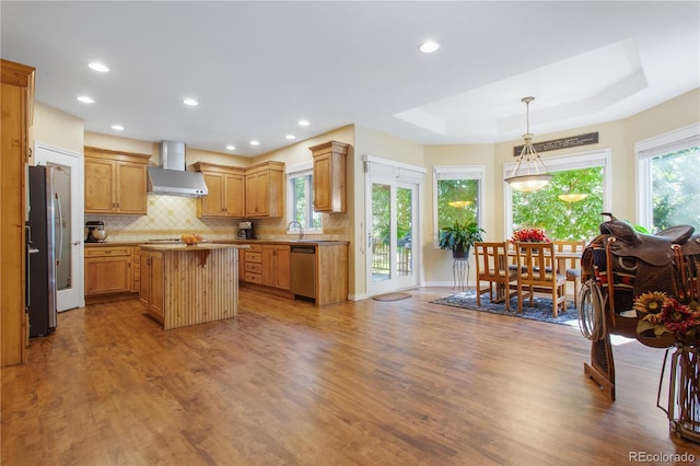 kitchen featuring a tray ceiling, backsplash, wood finished floors, stainless steel appliances, and wall chimney exhaust hood