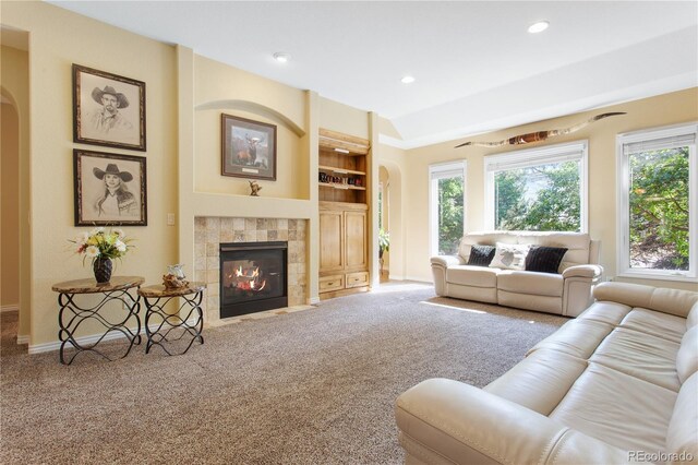 carpeted living room featuring a wealth of natural light, arched walkways, built in shelves, and a tiled fireplace