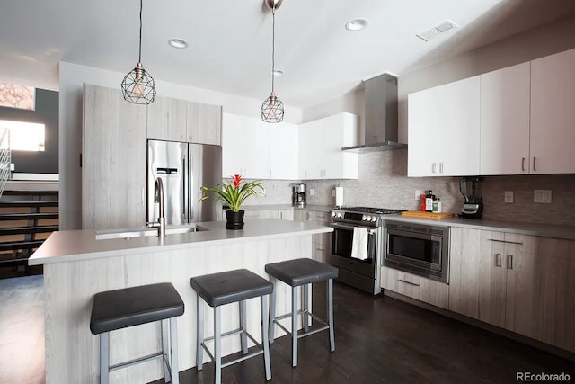 kitchen featuring an island with sink, hanging light fixtures, dark wood-type flooring, wall chimney range hood, and appliances with stainless steel finishes