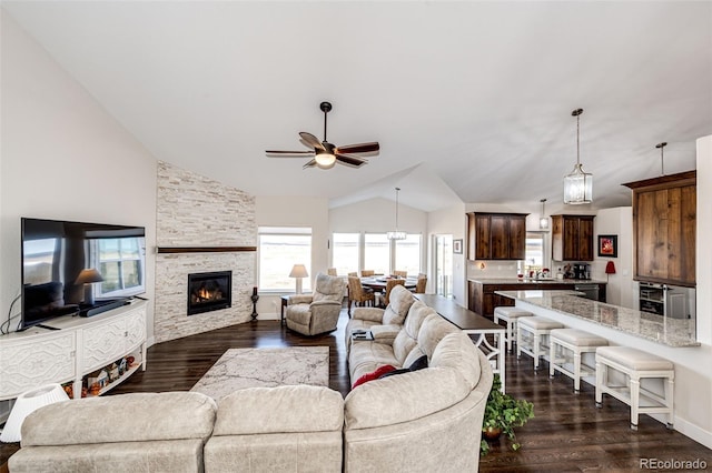 living room with a ceiling fan, dark wood finished floors, vaulted ceiling, and a stone fireplace