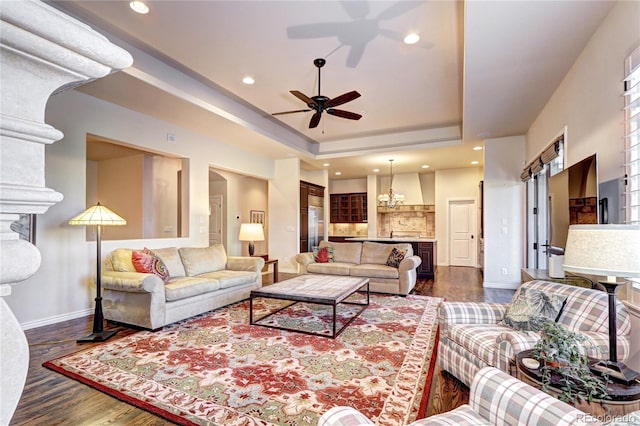 living room with recessed lighting, ceiling fan with notable chandelier, dark wood finished floors, baseboards, and a raised ceiling