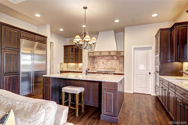 kitchen with stainless steel built in fridge, a sink, light stone countertops, glass insert cabinets, and custom range hood