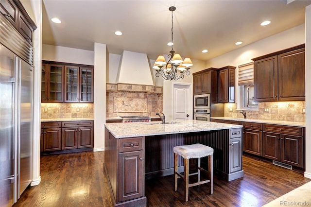 kitchen with custom range hood, glass insert cabinets, a kitchen island with sink, a sink, and built in appliances