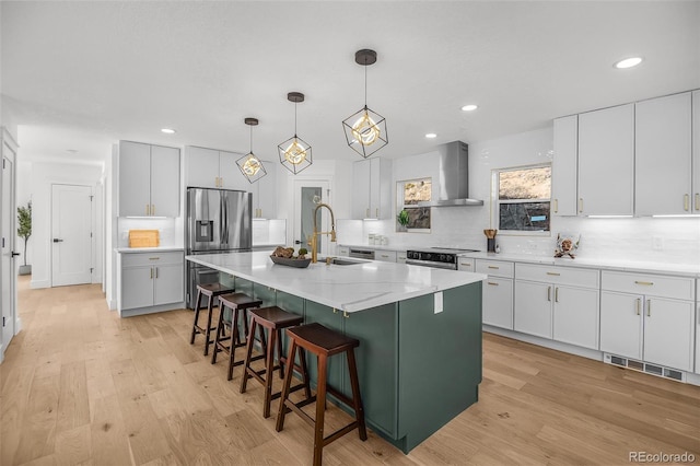 kitchen with pendant lighting, wall chimney range hood, sink, white cabinetry, and an island with sink