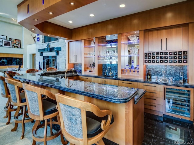 bar with dark stone countertops, tasteful backsplash, beverage cooler, and a stone fireplace