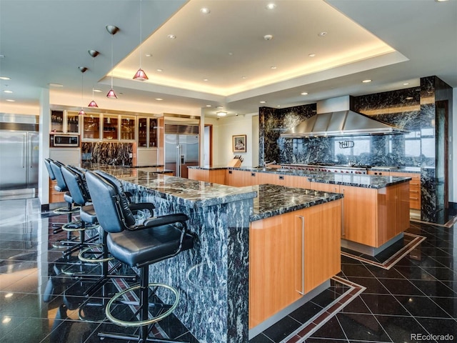 bar featuring built in appliances, a raised ceiling, decorative backsplash, wall chimney range hood, and dark stone counters