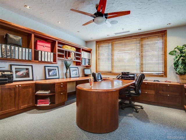 carpeted office space with a textured ceiling, ceiling fan, and built in desk