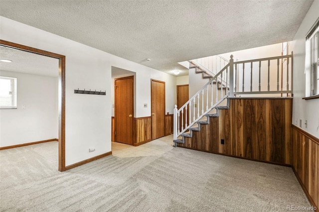 unfurnished living room with a textured ceiling, stairs, carpet, a wainscoted wall, and wooden walls