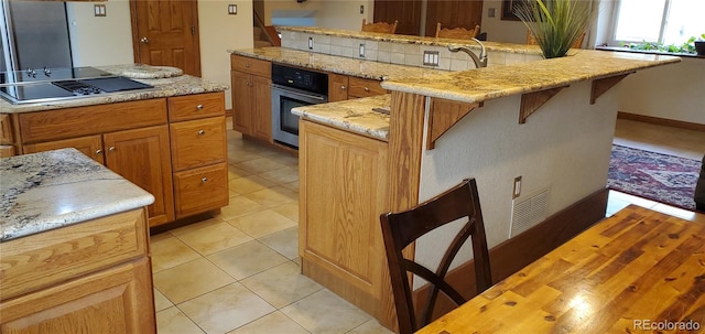 kitchen featuring black electric stovetop, stainless steel oven, a kitchen island, light tile patterned floors, and light stone counters