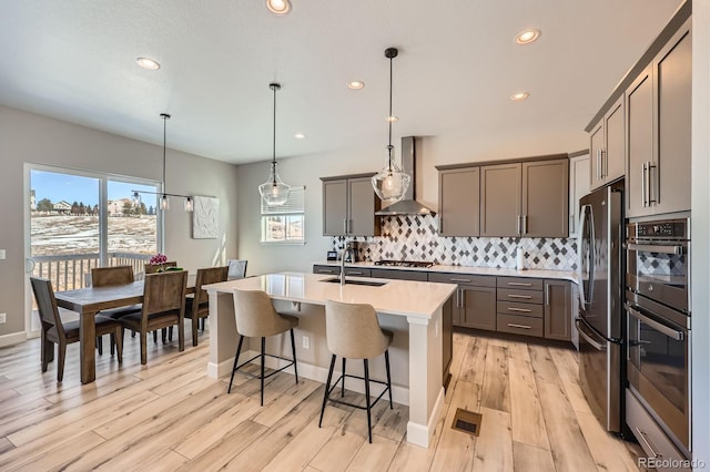 kitchen with gray cabinetry, stainless steel appliances, a sink, decorative backsplash, and wall chimney exhaust hood