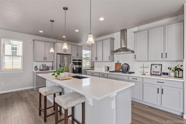 kitchen featuring wall chimney exhaust hood, gray cabinetry, stainless steel appliances, decorative backsplash, and a center island with sink