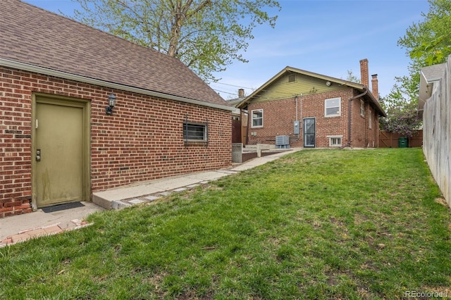 rear view of house with fence, brick siding, a yard, central AC unit, and a shingled roof