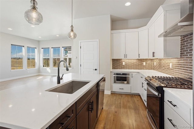 kitchen featuring appliances with stainless steel finishes, wall chimney range hood, white cabinetry, pendant lighting, and a sink