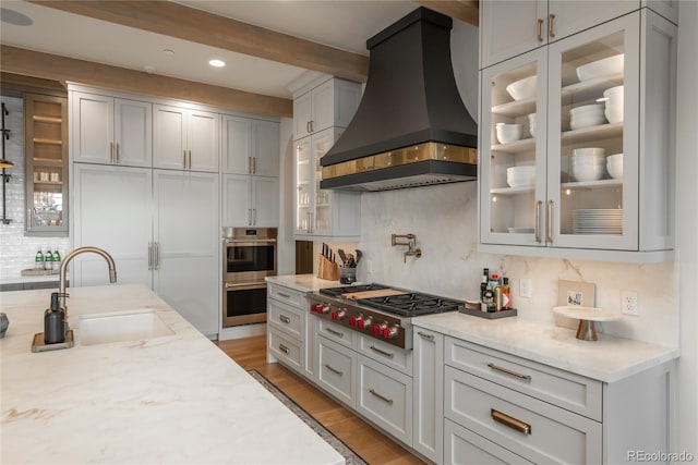 kitchen with light stone counters, custom range hood, backsplash, light wood-type flooring, and appliances with stainless steel finishes