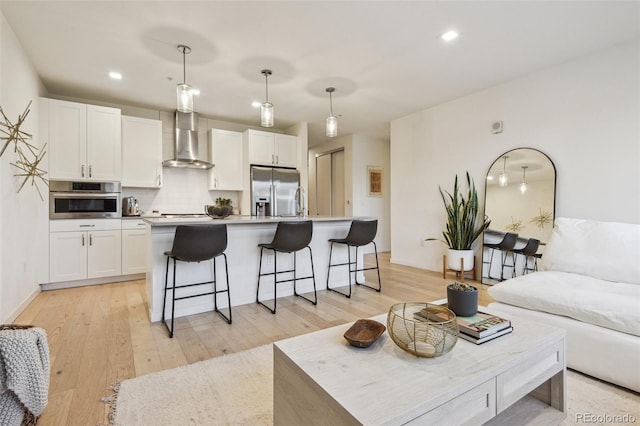 kitchen featuring appliances with stainless steel finishes, wall chimney range hood, pendant lighting, white cabinetry, and an island with sink