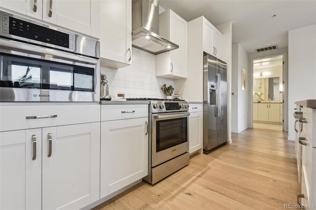 kitchen with decorative backsplash, appliances with stainless steel finishes, wall chimney range hood, light hardwood / wood-style flooring, and white cabinetry